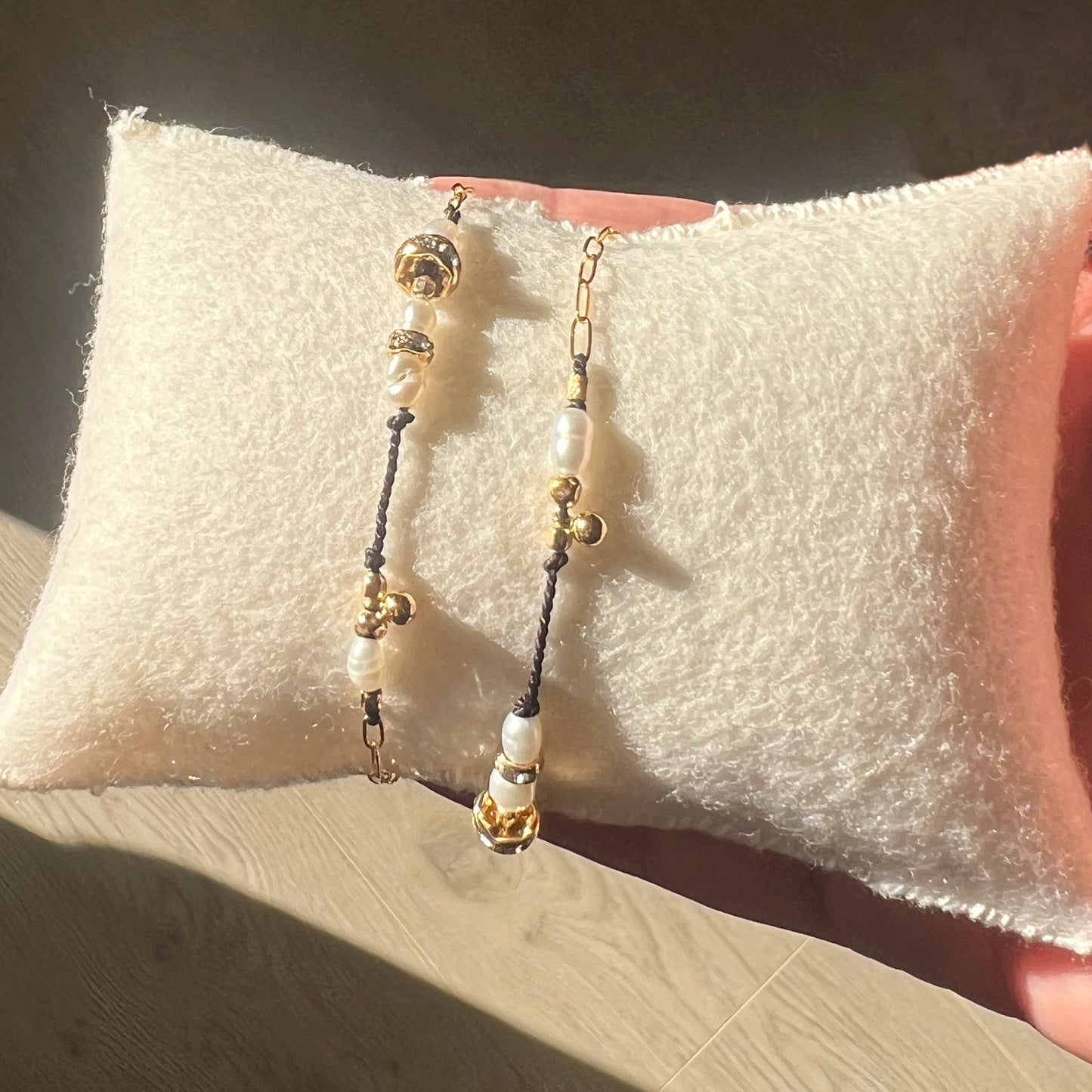 Two bracelets featuring silk thread with freshwater pearls and glass beads, including 14K gold filled elements. Displayed on a white cushion is sunlit room.