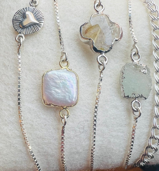 Detail of various silver slider bracelets with multiple charms including a heart-shaped pendant, with a dainty chain on left side of image.