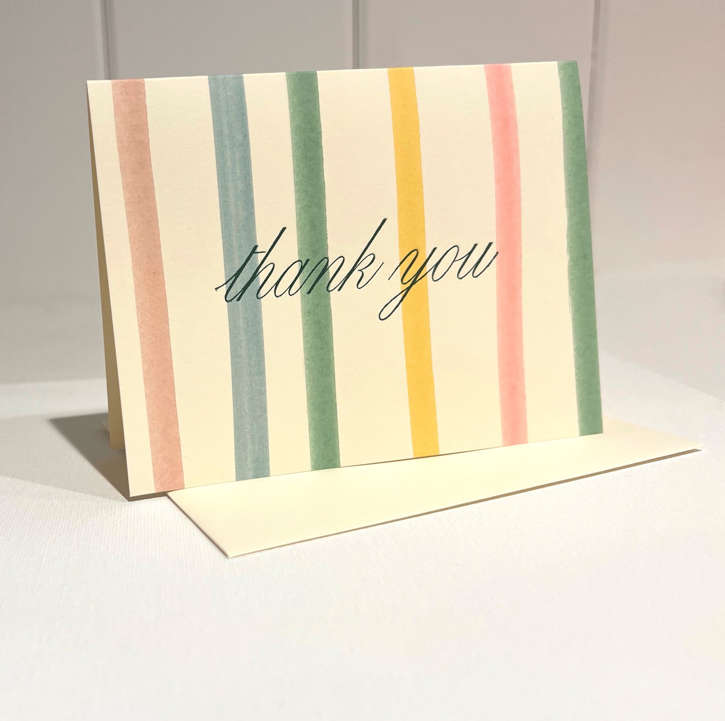 Colorful Stripe card with script 'thank you' foil text on a light background with off white envelope.
Card standing upright on envelope.