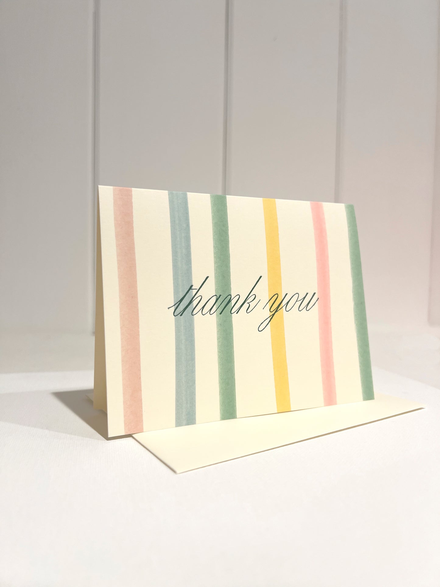 Pastel Stripe card with script 'thank you' foil text on a light background with off white envelope.
Card standing upright on envelope.