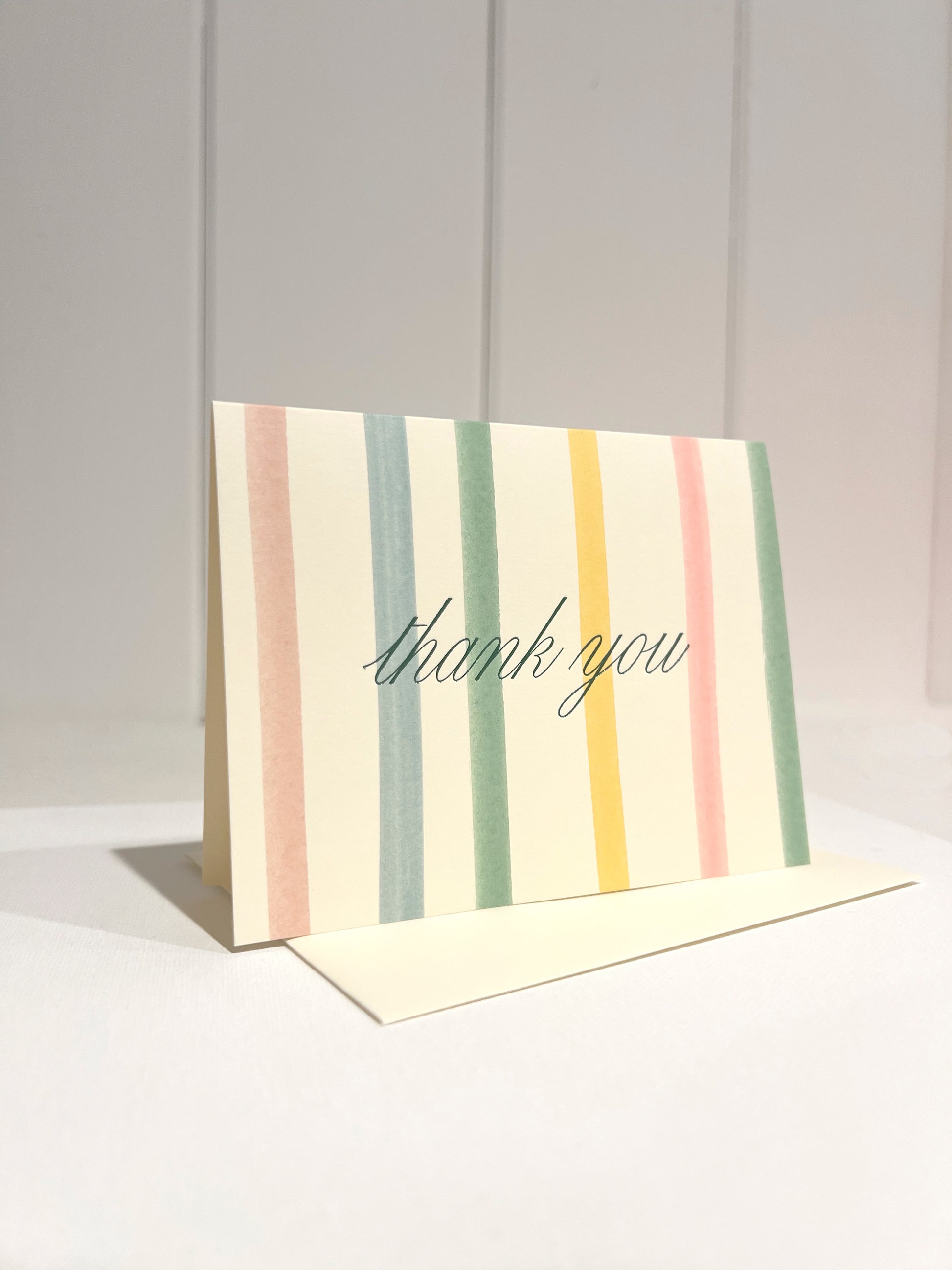 Pastel Stripe card with script 'thank you' foil text on a light background with off white envelope.
Card standing upright on envelope.