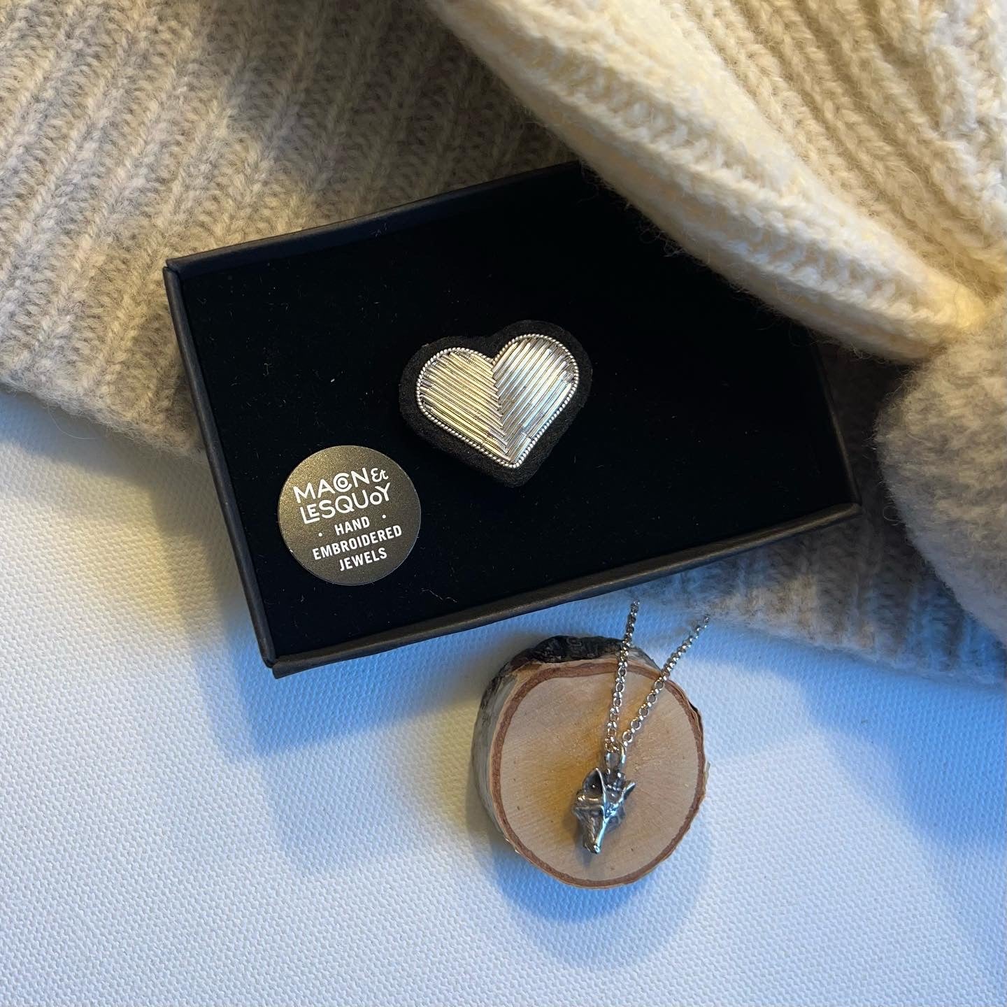 Macon and LesQuoy heart shape design brooch. Bullion silver color and thread embroidery on black felt backing in black packaging surrounded by knit hat and silver necklace for size comparison. each item sold separately